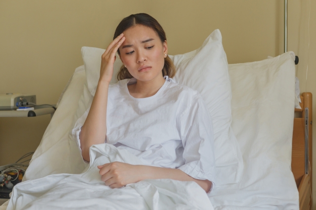 Woman in a hospital bed wearing a white gown, holding her head in discomfort, possibly experiencing a headache or pain.
